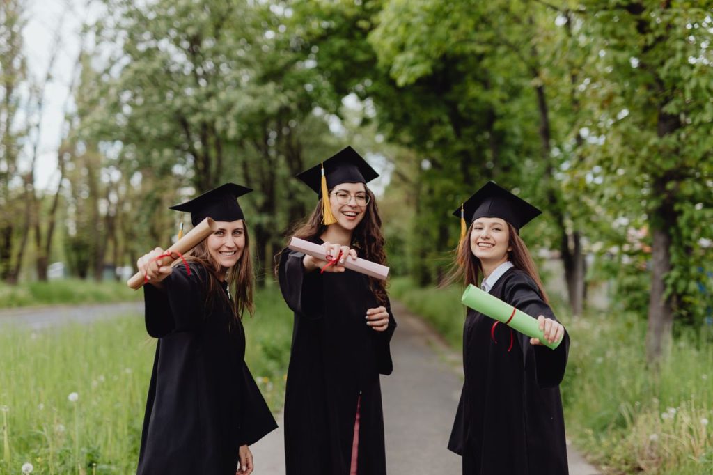 Young women in graduation gowns celebrating with diplomas in a park.